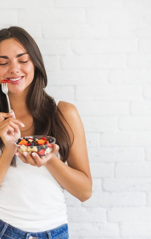 happy-young-woman-standing-against-white-wall-eating-fruit-salad